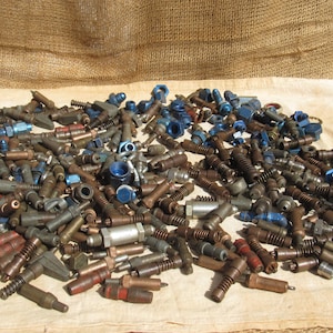 May include: A collection of various metal machine parts, including bolts, nuts, and fittings, in shades of brown, silver, and blue. The parts are scattered on a light-colored surface with a burlap backdrop.