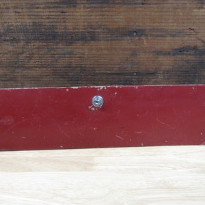 May include: A red metal box with a single screw in the center. The box is slightly worn and has a matte finish.
