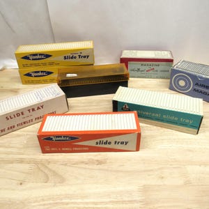 May include: A collection of vintage slide trays for storing and organizing photographic slides. The trays are made of cardboard and plastic and feature various brand names, including "Yankee", "Airequipt", and "Universal".