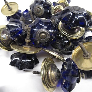 May include: A collection of faceted, dark blue glass cabinet knobs with brass-toned metal bases and screws. The knobs have a starburst design and are piled together, showcasing their decorative hardware style.