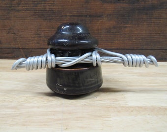 Op de afbeelding: Een bruine keramische isolator met een afgeronde boven- en onderkant, omwikkeld met zilverkleurige draad. De isolator heeft een glanzende afwerking en staat op een houten oppervlak. De draad is strak om de hals van de isolator gewikkeld.