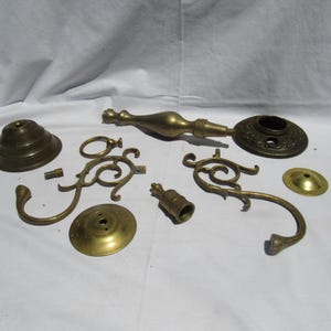 May include: A collection of disassembled brass components for a light fixture or lamp, including a tiered base, two ornate curved arms, a central spindle with a circular decorative plate, a small cylindrical connector, and two flat circular discs.