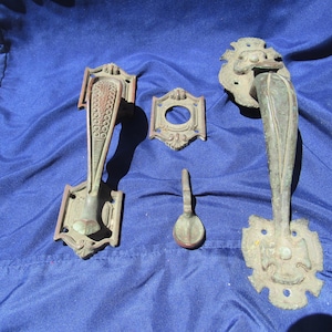 May include: A collection of antique bronze-toned metal hardware including two ornate door handles with intricate detailing, a square-shaped escutcheon plate with a central circular opening, and a small, curved metal latch component.