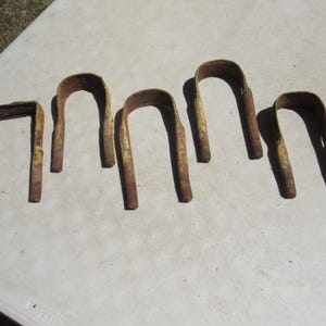 May include: Six rusty metal U-shaped brackets with yellow paint remnants. The brackets are arranged on a white surface. The metal has a weathered appearance, suggesting outdoor use or age. The brackets vary slightly in size.