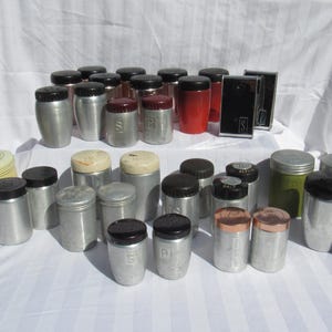May include: A collection of vintage salt and pepper shakers and spice containers. The assortment includes metal, red, green, and black containers with various lid styles. Some shakers have the letter "S" or "P" on them.