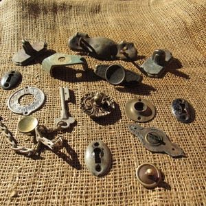 May include: A collection of antique hardware pieces, including keys, keyholes, and decorative metal accents. The items are in various shapes and sizes, with a weathered, aged appearance. The hardware is arranged on a textured, tan fabric.