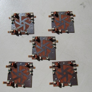May include: Five small, square, brown circuit boards with copper wiring and a starburst pattern in the center. The boards have small, white, rectangular components around the edges.