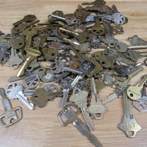 May include: A collection of various metal keys, in shades of silver and gold, scattered on a light brown wooden surface. The keys vary in size and design, with some showing stamped markings. The image presents a variety of key types.