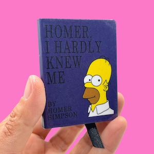 Puede incluir: Un pequeño libro rectangular con una cubierta azul oscuro. El texto dice "HOMER, I HARDLY KNEW ME" con una ilustración de Homer Simpson. El autor es Homer Simpson. El libro se sostiene sobre un fondo rosa.