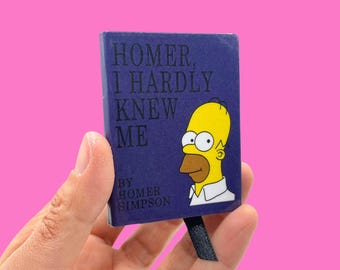 May include: A small, rectangular book with a dark blue cover featuring the text "HOMER, I HARDLY KNEW ME" and an illustration of Homer Simpson. The book is held against a pink background. The author is listed as Homer Simpson.