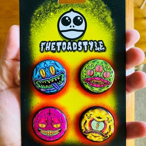 Puede incluir: Un conjunto de cuatro pines de esmalte de colores con caras de monstruos de dibujos animados. Los pines se muestran en una tarjeta negra con el texto "THETOADSTYLE".