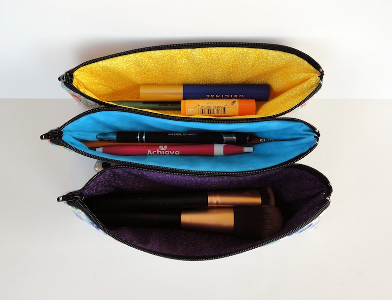 May include: Three zippered pouches with different colored linings. The top pouch is yellow and contains a blue and yellow lip balm and a black and silver lip gloss. The middle pouch is blue and contains a red and silver pen and a black and silver pen. The bottom pouch is purple and contains a black makeup brush.