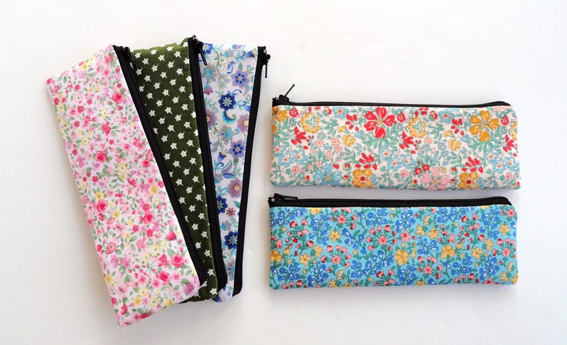 May include: Six zippered pouches with floral patterns. The pouches are in various colors, including pink, green, blue, and white. The pouches are lined with black fabric and have black zippers. The pouches are arranged in a row on a white surface.