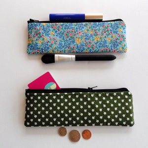 May include: Two fabric zippered pouches. The top pouch is blue with a floral pattern and has a black makeup brush and a blue tube of L'Oreal mascara inside. The bottom pouch is green with a white star pattern and has a pink card and three coins inside.