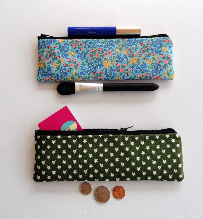 May include: Two fabric zippered pouches. The top pouch is blue with a floral pattern and has a black makeup brush and a blue tube of L'Oreal mascara inside. The bottom pouch is green with a white star pattern and has a pink card and three coins inside.