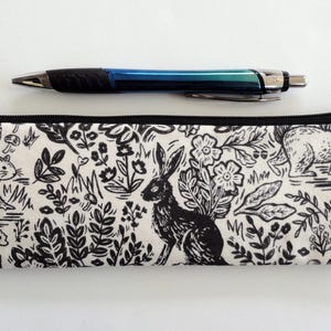 May include: A black and white pencil case with a woodland animal print, featuring a rabbit, bear, and squirrel. A blue and black pen rests on top of the zippered case.