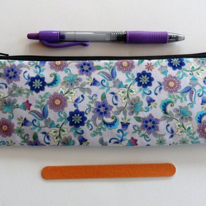 May include: A white zippered pencil case with a floral pattern in shades of blue, purple, and gold. The case is lying flat on a white surface.