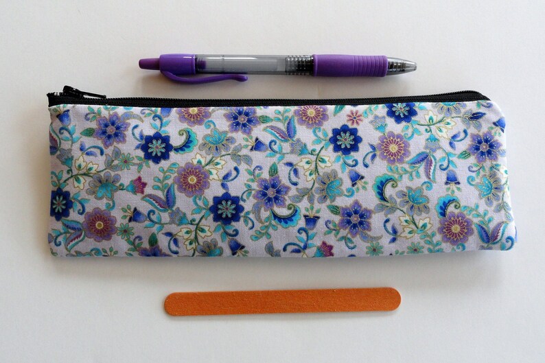 May include: A white zippered pencil case with a floral pattern in shades of blue, purple, and gold. The case is lying flat on a white surface.