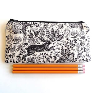 May include: A rectangular pencil case with a black zipper, featuring a black and white forest scene with a running hare, squirrels, and foliage. Three orange pencils with pink erasers are below the case.