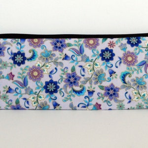 May include: A lavender zippered pouch with a floral pattern in shades of blue, purple, and gold. The pouch is made of fabric and has a black zipper.