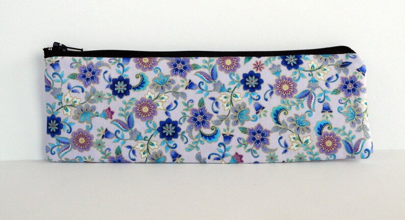 May include: A lavender zippered pouch with a floral pattern in shades of blue, purple, and gold. The pouch is made of fabric and has a black zipper.