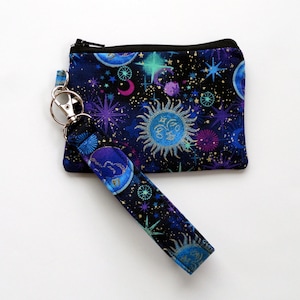 May include: A celestial-themed coin purse and wristlet set. The navy blue fabric features a cosmic design with suns, moons, stars, and planets in shades of blue, purple, and teal. The wristlet strap is attached with a silver-tone clasp.