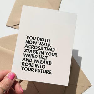 Pode incluir: Um cartão de felicitações creme com texto preto: "YOU DID IT! NOW WALK ACROSS THAT STAGE IN YOUR WEIRD HAT AND WIZARD ROBE INTO YOUR FUTURE." O cartão está sobre uma superfície branca com envelopes castanhos.