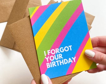 May include: A greeting card with the text "I FORGOT YOUR BIRTHDAY" in white, set against a colorful diagonal striped background of blue, green, yellow, and pink. The card is held with a stack of brown envelopes.