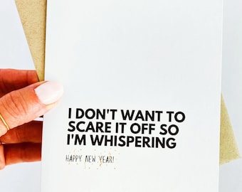 May include: A white card with black text that reads "I don't want to scare it off so I'm whispering Happy New Year!" The card is held in a hand with a gold ring.