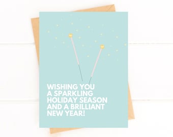 May include: A light blue card with a white border and gold stars. The card has a pink and white illustration of two sparklers and the text "Wishing you a sparkling holiday season and a brilliant new year!"