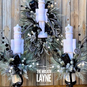 May include: Three white nutcracker figurines with black and white checkered ribbon and black and white ornaments on white pedestals. The text "THE WREATH LAYNE" is visible in the center of the image.