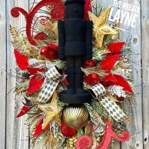 May include: A festive Christmas wreath featuring a black nutcracker figure, red poinsettias, gold stars, and decorative ribbons. The wreath is adorned with gold and red glitter accents, creating a holiday display.