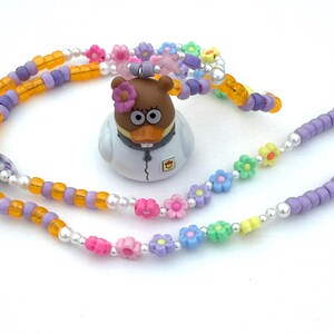 May include: A beaded necklace with a variety of colorful beads, including orange, purple, and white. The necklace features a cartoon duck figurine with a flower on its head. The necklace is adorned with flower-shaped beads.