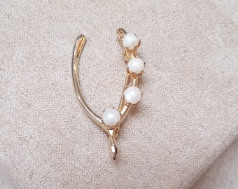 Puede incluir: Broche dorado en forma de horquilla, con una ramita de cuatro perlas blancas. El broche se encuentra sobre un fondo neutro y texturizado. El diseño es elegante y adecuado para diversas ocasiones.