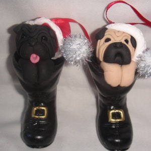 Könnte beinhalten: Zwei Weihnachtsbaumschmuckstücke mit Mops-Hunden, die Weihnachtsmützen tragen. Die Möpse schauen aus schwarzen Stiefeln mit goldenen Schnallen. Der Schmuck ist mit silbernem Glitzer verziert.