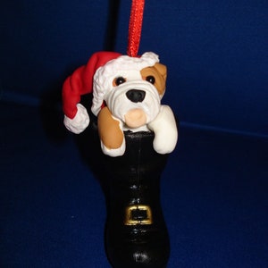 Könnte beinhalten: Ein Weihnachtsbaumschmuck mit einem weißen und braunen Bulldoggen, der einen roten Weihnachtsmann-Hut trägt und in einem schwarzen Stiefel mit einer goldenen Schnalle sitzt.