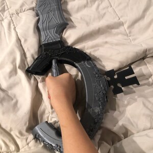 May include: A gray 3D printed sword with intricate details and a circular guard. The sword is held by a hand.