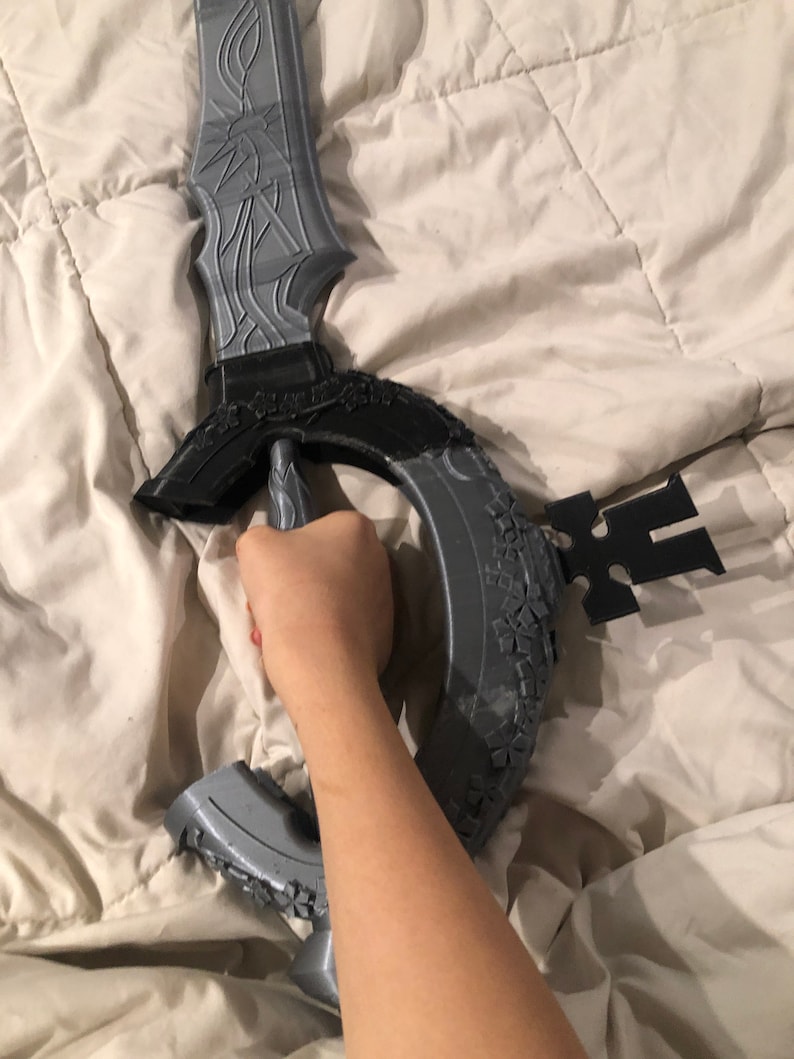 May include: A gray 3D printed sword with intricate details and a circular guard. The sword is held by a hand.