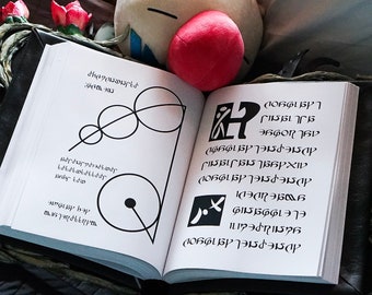 May include: An open book with white pages, featuring black text and geometric designs. The book is bound in a dark cover and rests on a surface with decorative elements. A red sphere and a plush toy are visible in the background.