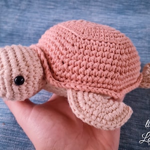 Op de afbeelding: Een gehaakt schildpad speelgoed in roze en beige. De schildpad heeft een kleine zwarte knoop voor een oog.
