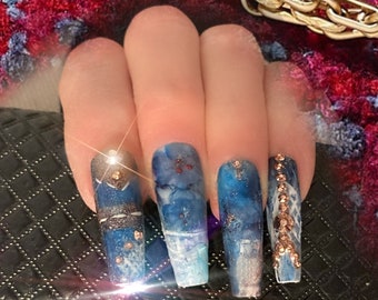 Puede incluir: Primer plano de uñas largas, azules y blancas con purpurina dorada y pedrería. Las uñas están pintadas con un efecto de mármol.