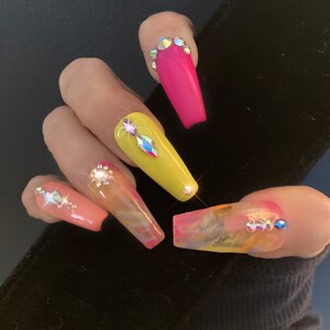 May include: Close-up of a hand with long, colorful, and decorated acrylic nails. The nails are painted in shades of pink, yellow, and clear with a marble design. Each nail is adorned with rhinestones.