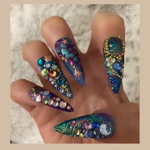 May include: Close-up of five long, pointed, blue and purple nail tips decorated with colorful rhinestones, glitter, and gold accents. The nails are styled in a mermaid theme.