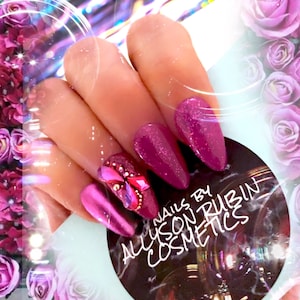 May include: Close-up of pink and purple glitter nail polish with rhinestones and a butterfly design. The nails are long and pointed. The image includes the text "Nails by Allyson Rubin Cosmetics".