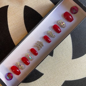 May include: A set of red press-on nails with a glossy finish, arranged in a black box. Some nails are adorned with sparkling rhinestones. Two iridescent gems are also included.