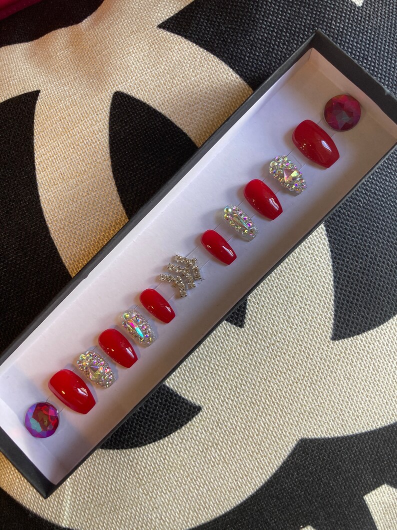May include: A set of red press-on nails with a glossy finish, arranged in a black box. Some nails are adorned with sparkling rhinestones. Two iridescent gems are also included.