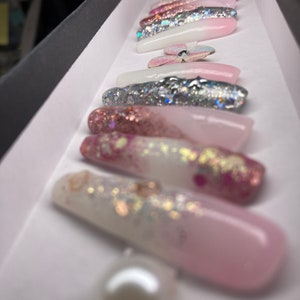 May include: A set of long, pink and white press-on nails with glitter and a pearl accent. The nails have a gradient design with pink fading to white.