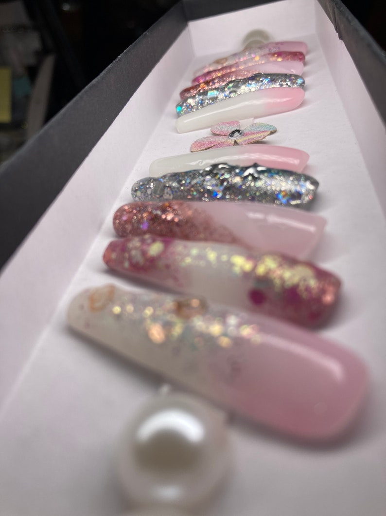 May include: A set of long, pink and white press-on nails with glitter and a pearl accent. The nails have a gradient design with pink fading to white.