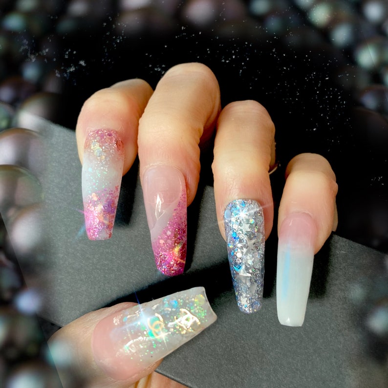 May include: Close-up of a hand with five long, clear acrylic nails. The nails are decorated with glitter and ombre designs in pink, white, and silver.