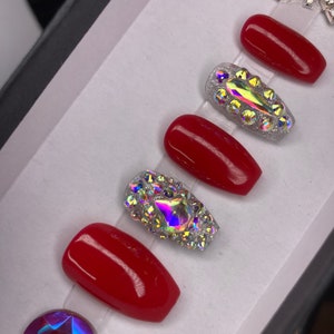 May include: A set of red press-on nails with two accent nails. The accent nails are covered in iridescent rhinestones. A large, faceted, iridescent gem is at the bottom left. Additional rhinestones are at the top right.